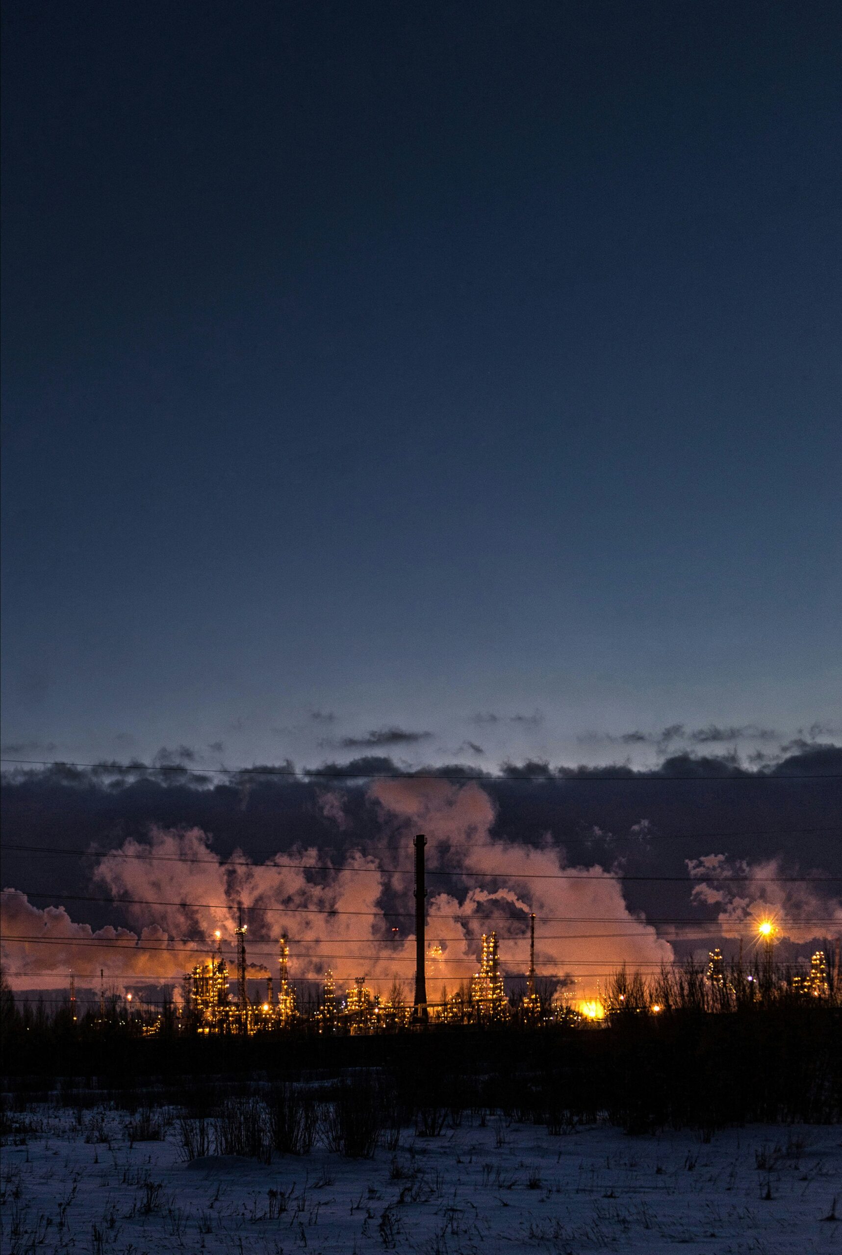 A fossil fuel refinery lit up at night under a dark sky with the city lights glowing over the horizon, image by anatoliy-shostak-unsplash